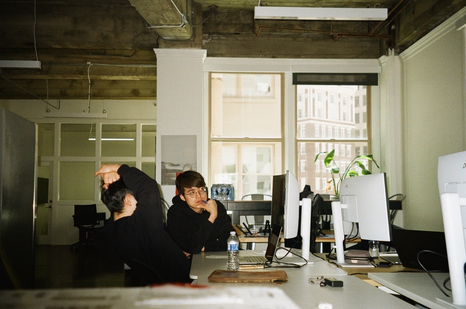 A loft office with tall windows. One person stretches at a desk while another looks at the camera.