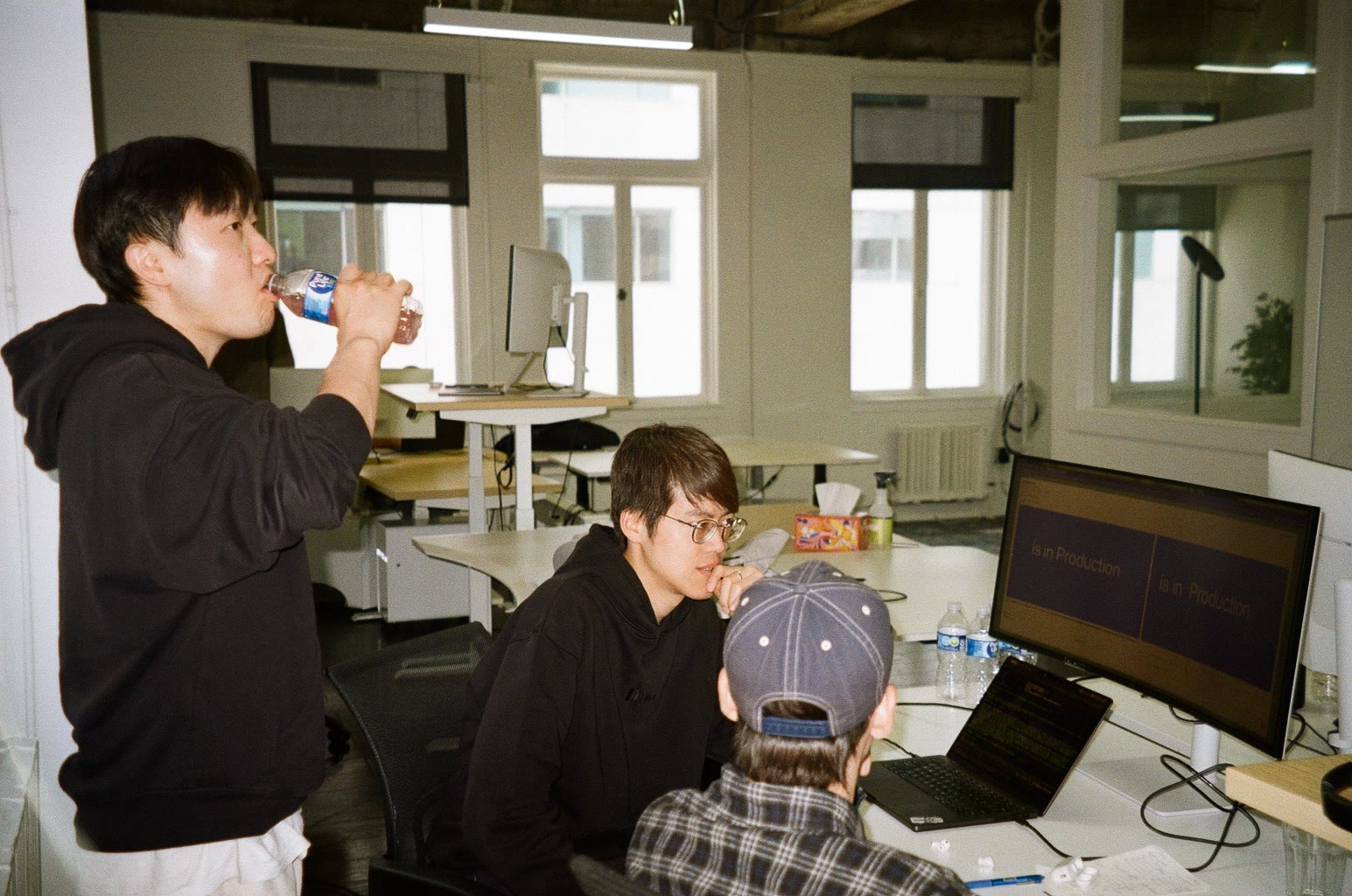 Three people at their desks, one drinking from a bottle, screens on